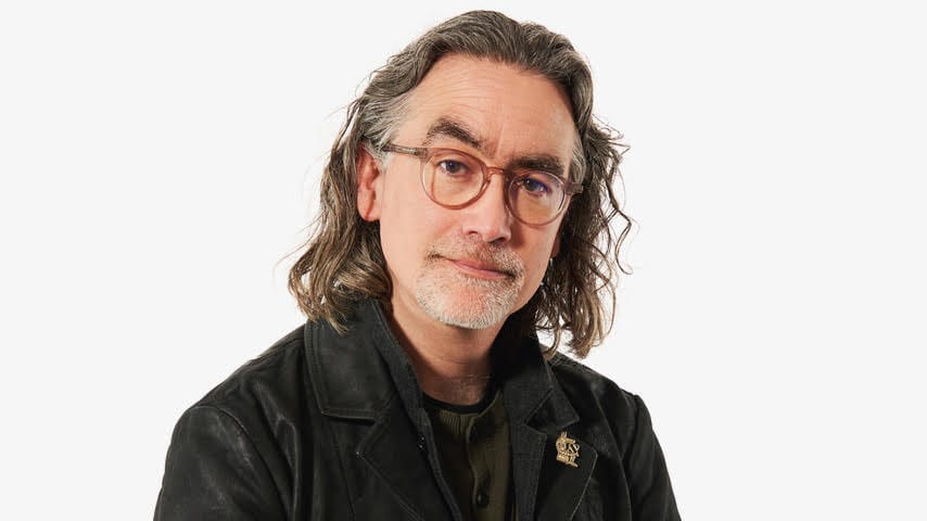 Headshot of a man with shoulder-length hair and glasses. Black shirt and jacket.