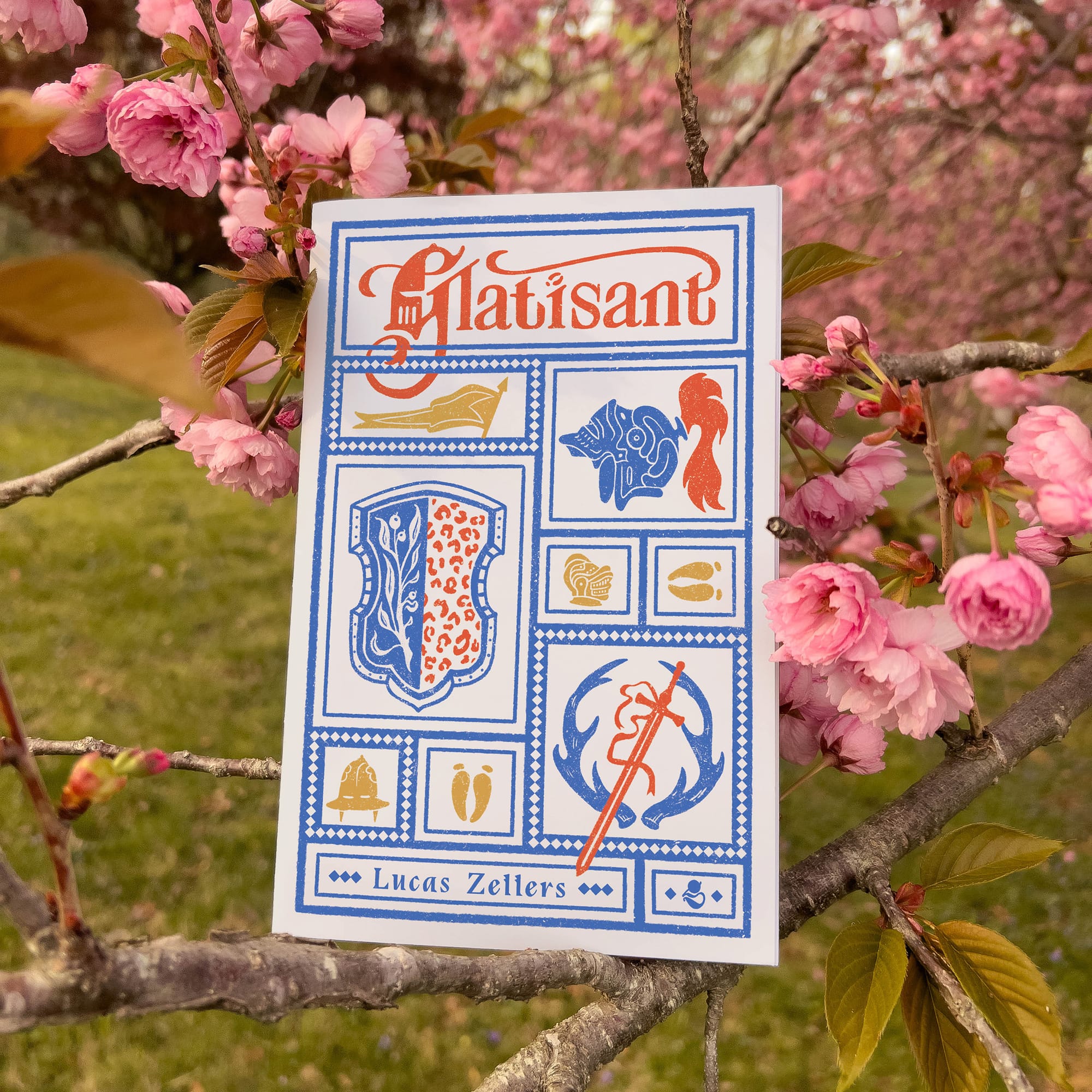 A photo of the Glatisant zine resting on the branches of a blooming cherry blossom tree. The cover for the Glatisant zine is a series of boxes with blue borders, inside are various arthurian objects like helmets, and hoof prints. 