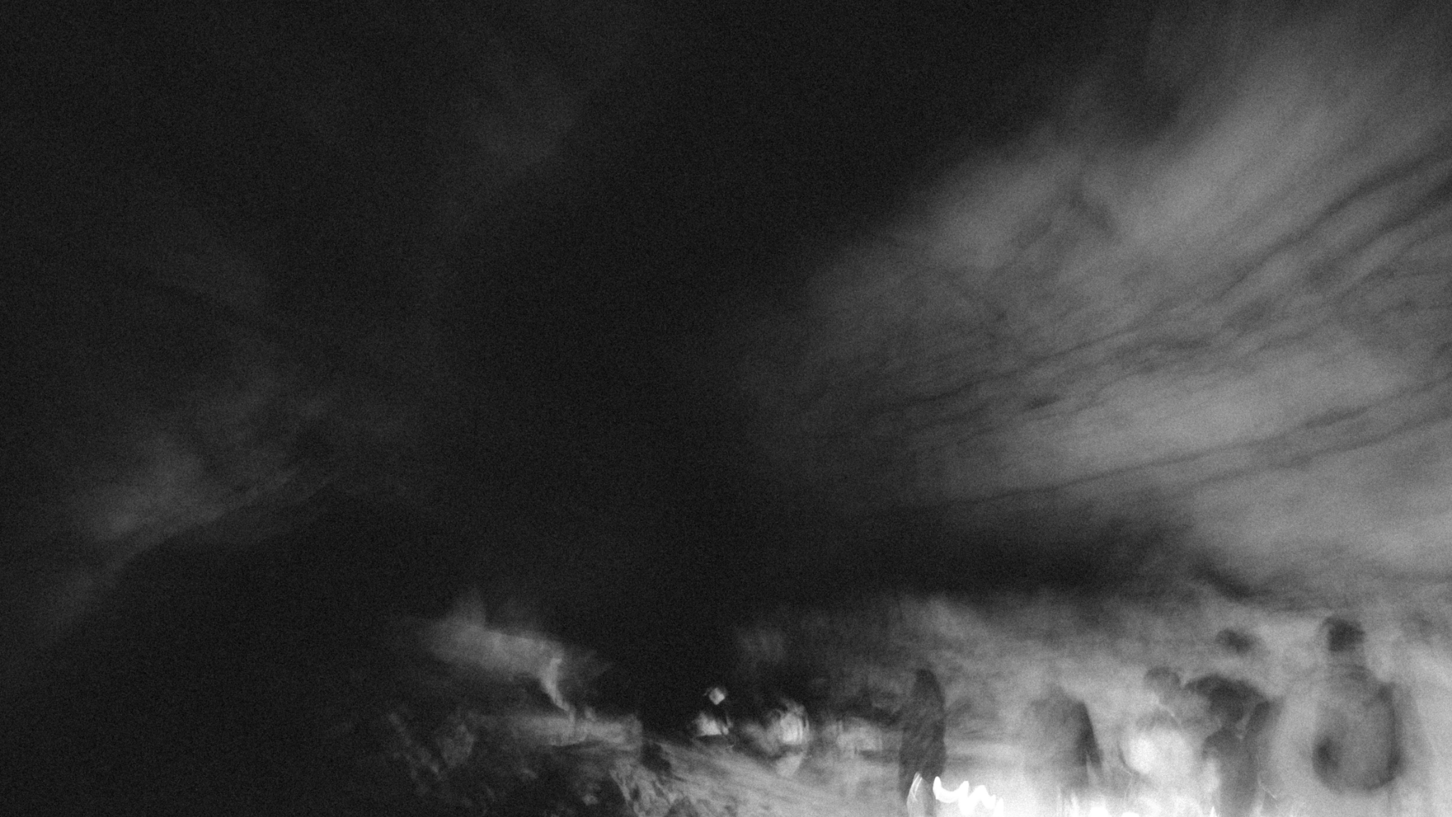 A long exposure photograph in black and white of a group of cavers walking through a cavern, with a ghostly doubling effect and low lighting creating a moody and dark image.