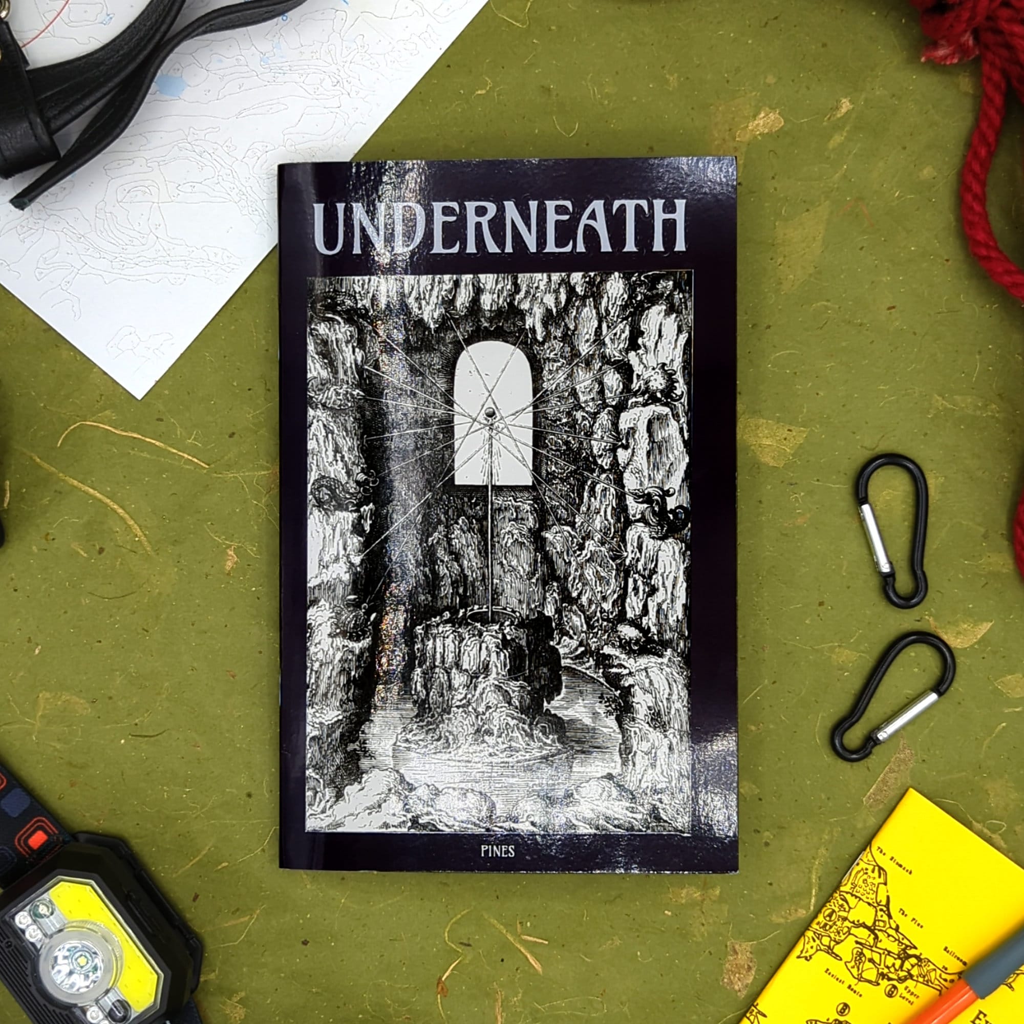 A photo of Underneath, showing the title, the author's name (Pines), and the stark black and white art on the cover, showing a geometric array of rope connecting together in a craggy cavern with water rising. The background of the image is green, and there's a map, carabiners, a headlamp, and rope visible.