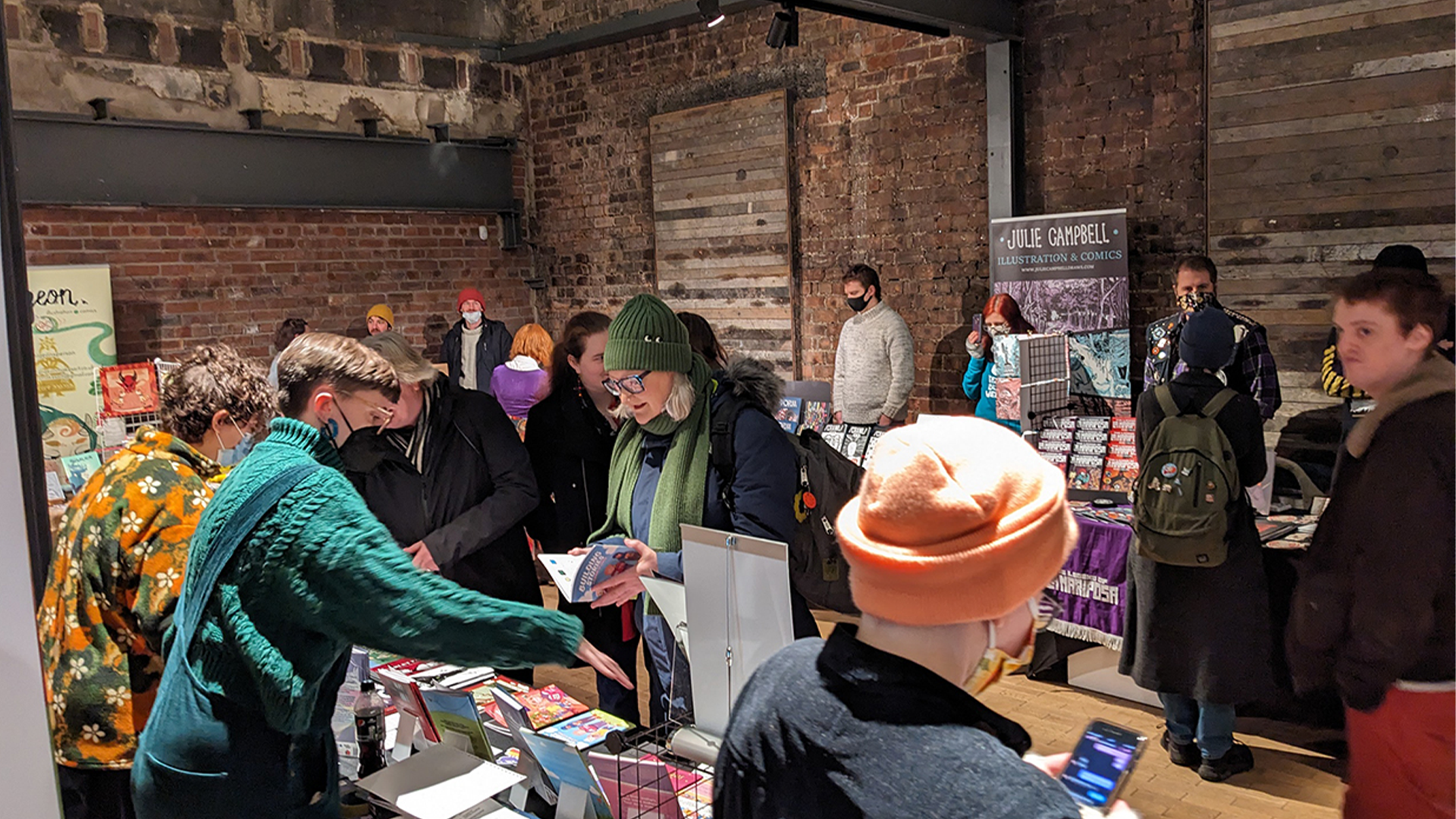 Photo of an industrial warehouse space, filled with merchants and public punters picking up, discussing and selling zines, comics, and books. Many attendees are wearing face masks.