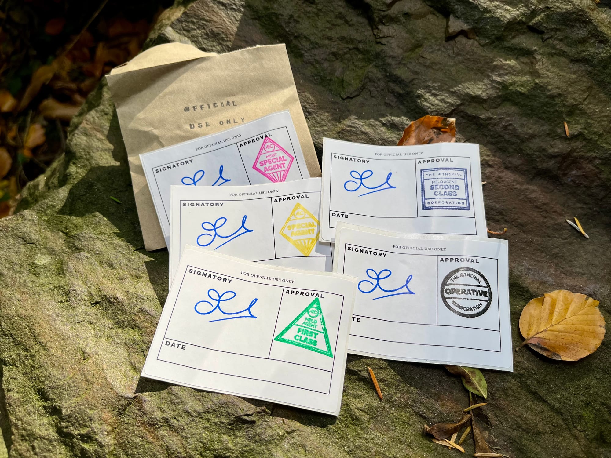 Five stickers lying on a rock with a brown square envelope with the words Official Use Only stamped on it. Each sticker has a signature and a colourful stamp with one of the five ranks on it and a box labelled 'Date'