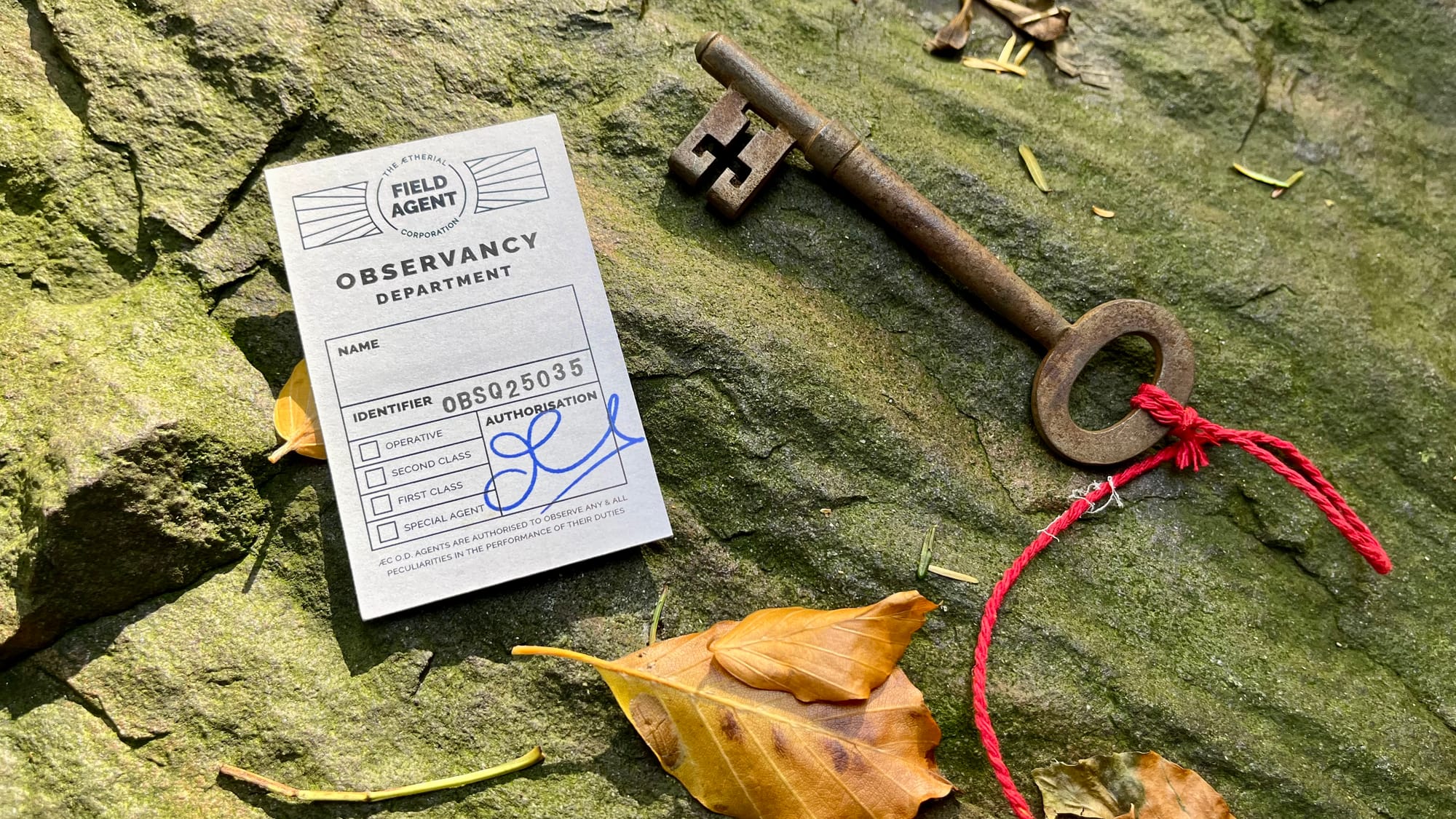 A Rusty key tied to a piece of red string next to a business card sized Field Agent ID card with space for a name, tick boxes to show rank and a unique identification number (OBSQ25035) and a signature under the heading' Authorisation'