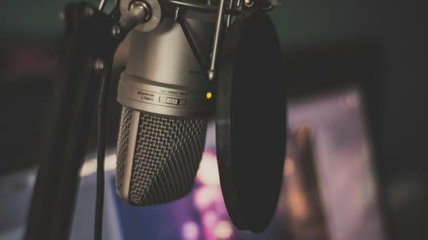 Photograph of a microphone and pop filter on a desk arm. A computer monitor in the background shows a blurred display