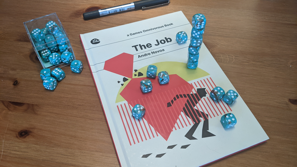 RPG book The Job sitting on a wooden table, surrounded by green d6 dice and a pen.