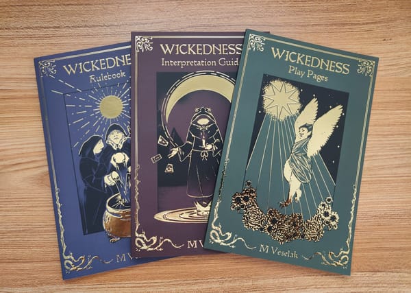 Three zines on a wooden desk, all titled Wickedness. From L to R, blue rulebook, red guide, and green play pages.