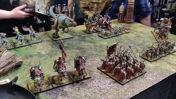 Two miniature armies on a green and brown table. One is knights in red capes, the other is mounted dinosaurs.