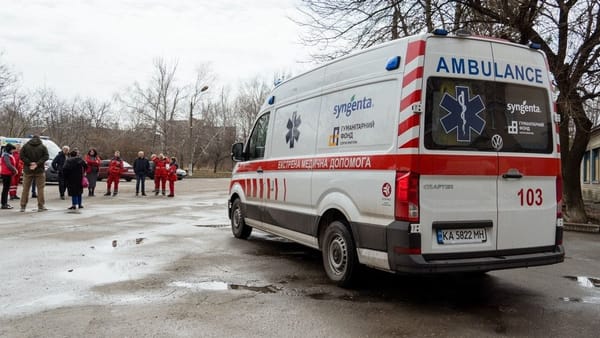 Ukrainian ambulance on ashphault. Roughly 12 people in red coats in middle ground. Cloudy day and apartment blocks in background.