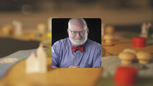 Headshot of John Stacy in blue collared shirt and red bow tie. Background is Spirit Island board game on table.